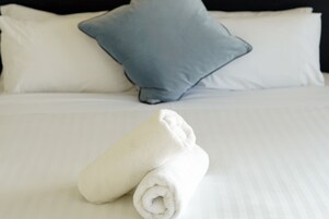 Cama bem arrumada com roupa de cama branca e toalha branca enrolada.