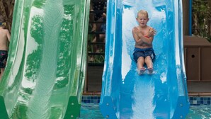 A water slide with two separate lanes, one green and one blue, in a pool area surrounded by wooden structures and greenery.