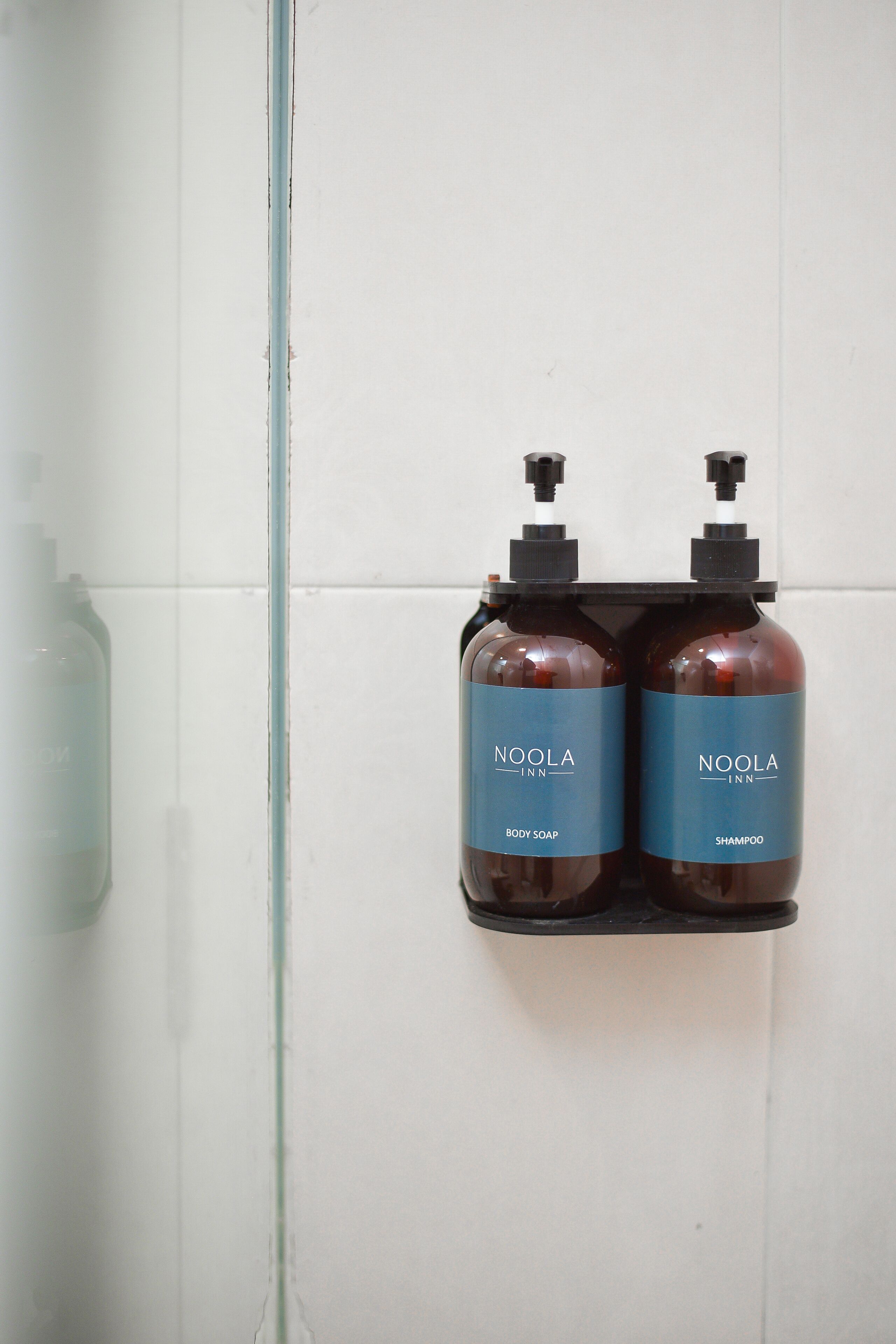 Dois frascos de sabonete e shampoo Noola Inn fixados em uma parede de azulejos brancos.