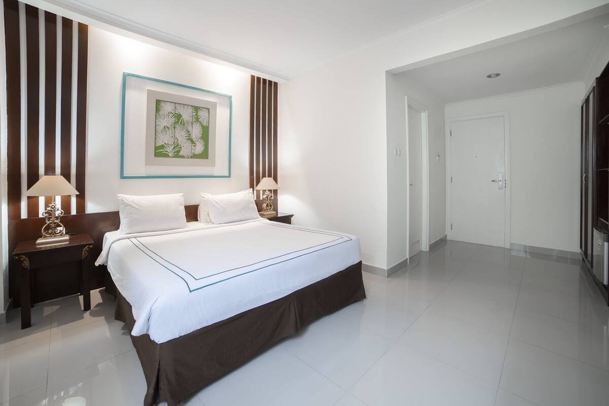 A hotel room with a large bed, two bedside tables with lamps, a framed artwork on the wall, and a door leading to another room.