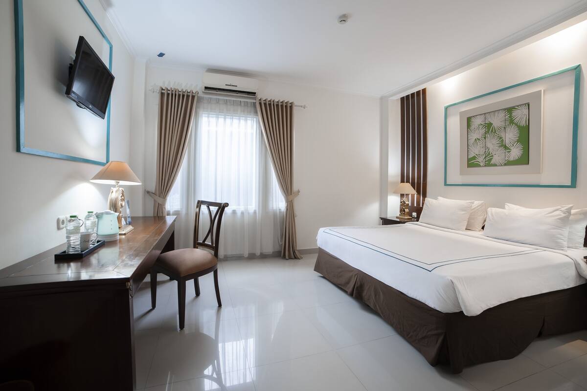 A hotel room with a bed, desk, chair, TV, and artwork.