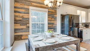 A dining area with a wooden table set for six, a large window, and a kitchen with white cabinets and a stainless steel refrigerator. - Hammocks & Forest Views: Creekside Murphy Retreat (Murphy)