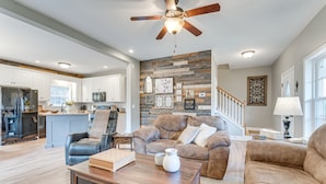 A spacious living room with a wooden coffee table, a brown leather sofa, and a blue leather chair. A kitchen with white cabinets and a dining area with a table and chairs are visible in the background. - Hammocks & Forest Views: Creekside Murphy Retreat (Murphy)