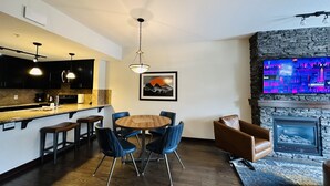 A modern kitchen with dark cabinets, a central island, and pendant lighting. There is a dining area with a round table and blue chairs. A fireplace with a stone surround is visible, and a flat-screen TV is mounted above it. - Mountain Retreat - Heated Pool & Hot-tub & Sauna (Canmore)