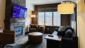 A living room with a stone fireplace, a television mounted on the wall, a sofa, a coffee table, and a large window with a view of a snowy landscape. - Mountain Retreat - Heated Pool & Hot-tub & Sauna (Canmore)