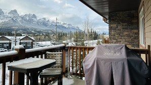 A balcony with a view of snow-covered mountains, a barbecue grill, and a wooden deck with a table and chairs. - Mountain Retreat - Heated Pool & Hot-tub & Sauna (Canmore)