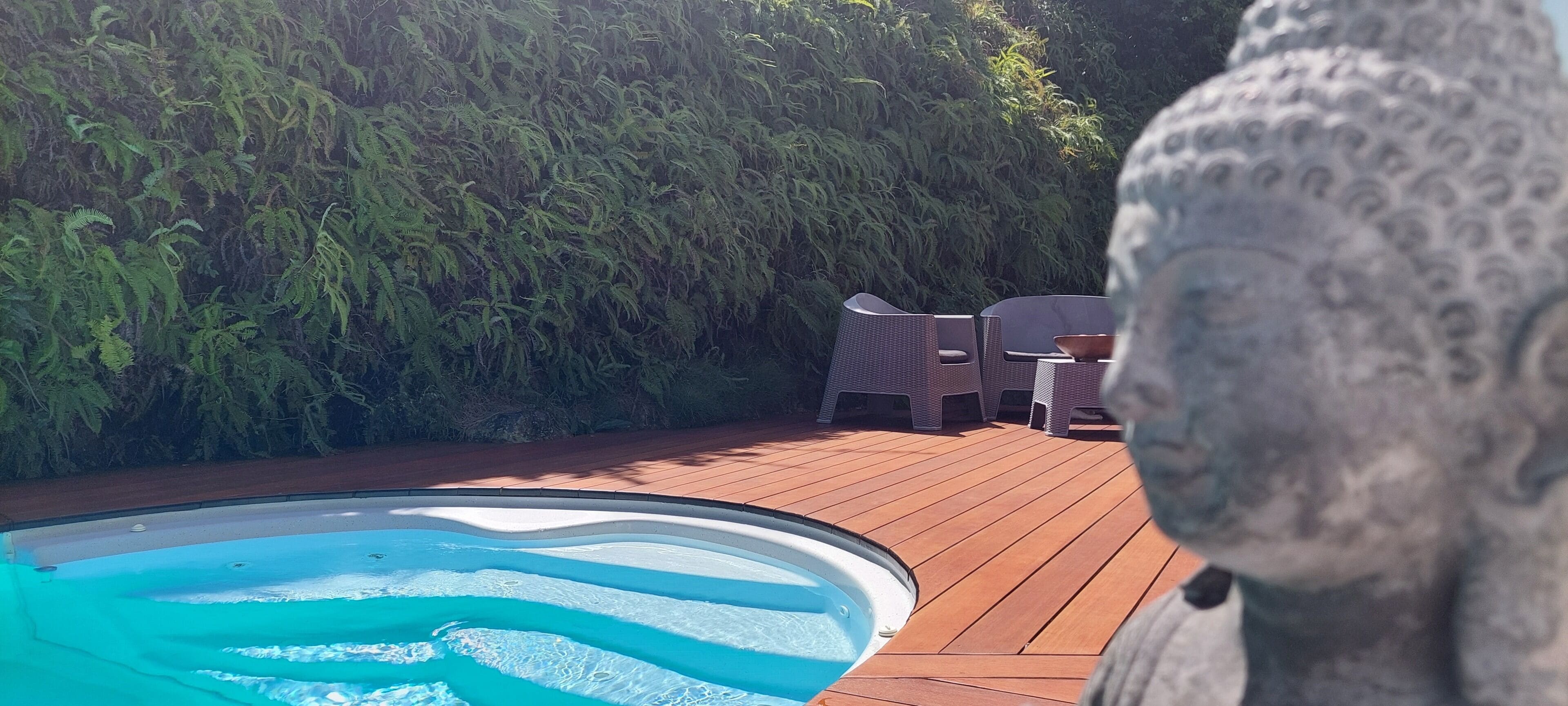 Une piscine dotée d’une terrasse en bois, entourée d’une végétation luxuriante et ornée d’une statue en pierre.