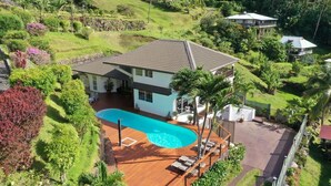 Una casa de dos pisos con piscina, rodeada de abundante follaje y palmeras.