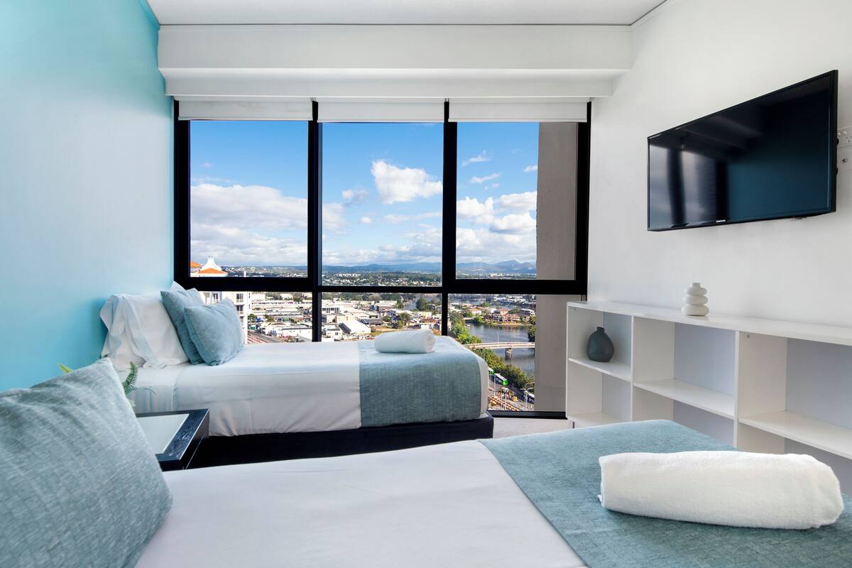 Une chambre d’hôtel moderne dotée d’une grande fenêtre donnant sur la ville, d’un téléviseur à écran plat fixé au mur et d’un lit soigneusement fait, aux couleurs bleu et blanc.