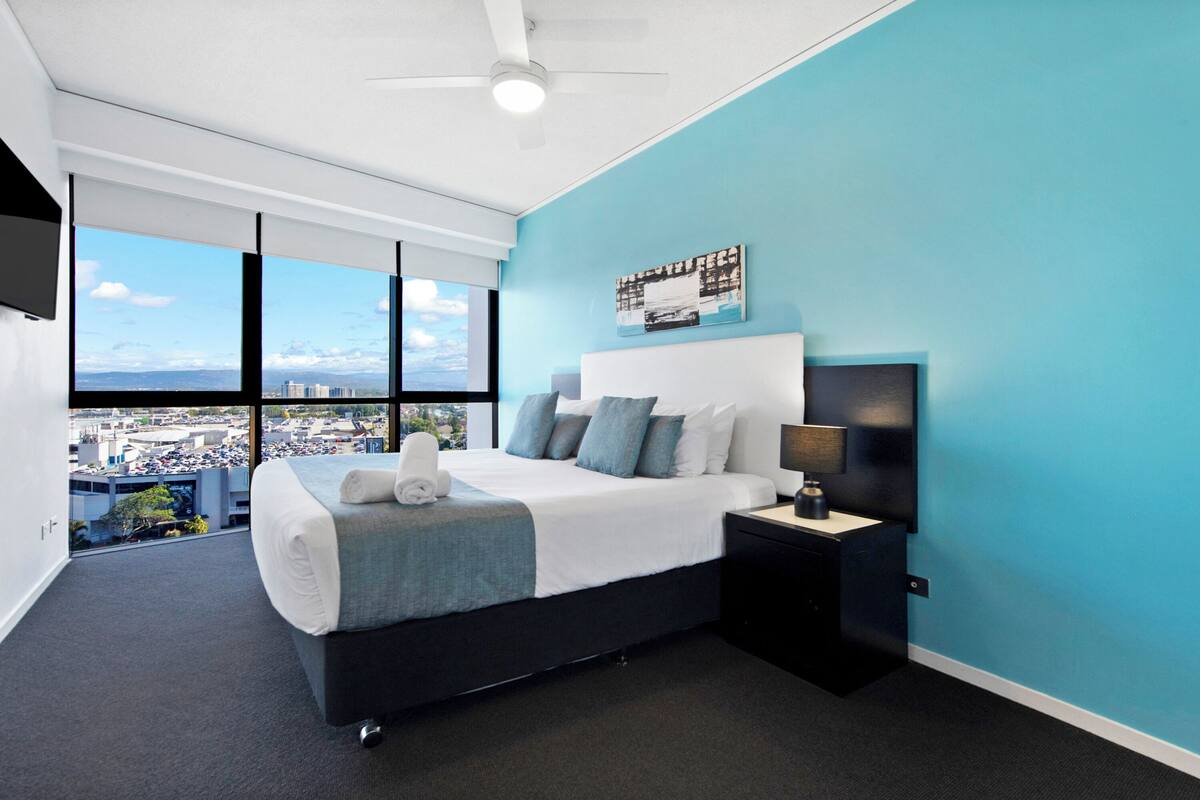 Une chambre moderne avec un grand lit, une table de chevet, un ventilateur de plafond et une vue sur la ville grâce à de grandes fenêtres.