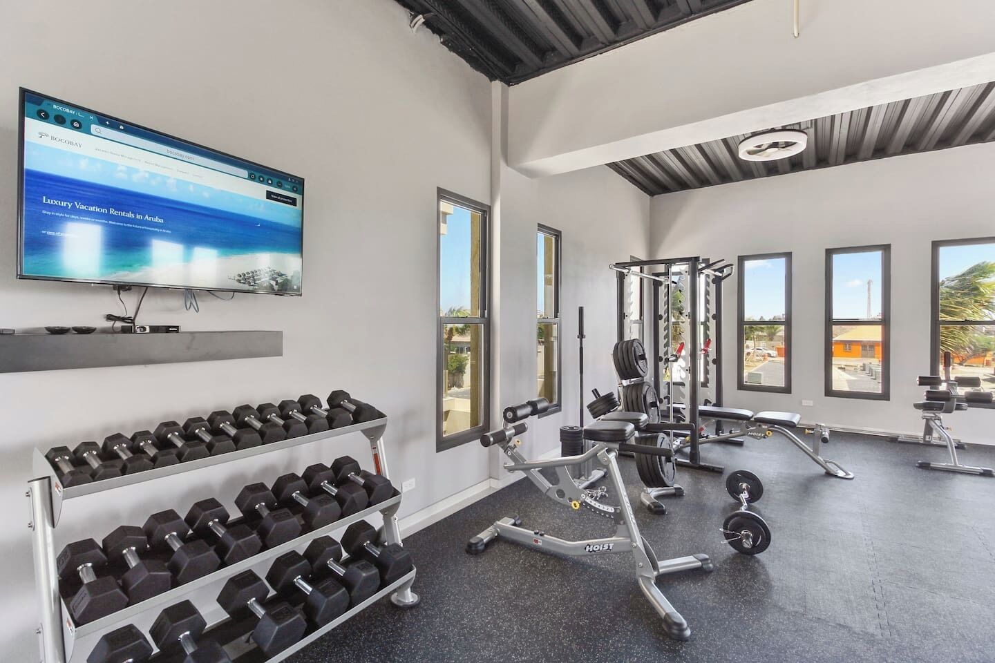 A gym with dumbbells, weight machines, and a TV displaying vacation rentals in Aruba.
