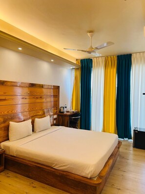 A hotel room with a wooden headboard, a large bed, a desk with a chair, and a ceiling fan. - Mak Hotel (Bengaluru)