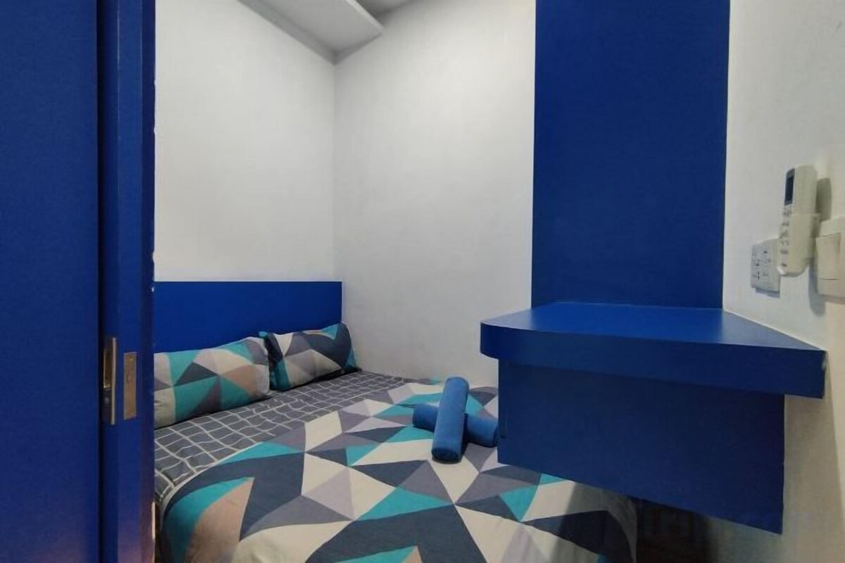 Een kleine, compacte kamer met een bed, een blauw hoofdbord, een nachtkastje en een aan de muur bevestigd telefoon.