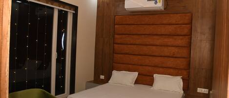 Habitación de hotel con cama, ventana con luces de cuerda, una silla verde y cabecero de madera.