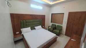 A bedroom with a bed, two green chairs, a window with a blind, a wooden door, and recessed ceiling lighting. - Hotel Govardhan Inn (Mathura)