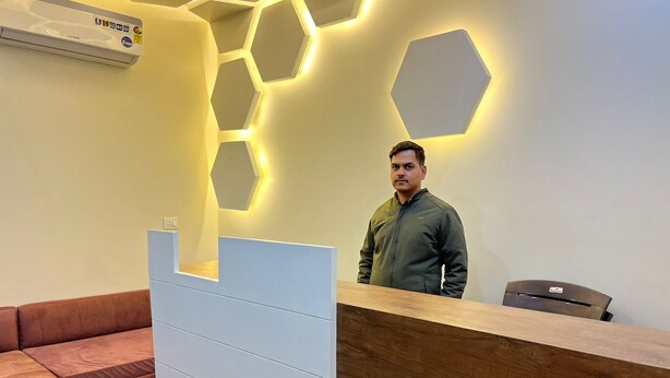 A reception area with a wooden reception desk, a brown leather couch, and a hexagonal light fixture on the wall. - Hotel Govardhan Inn (Mathura)