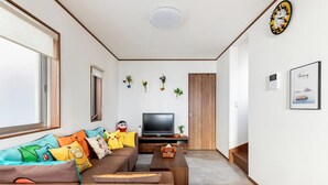 Un salón con un sofá marrón, un televisor de pantalla plana y una mesa de centro. Hay almohadones decorativos y una planta pequeña en el sofá.
