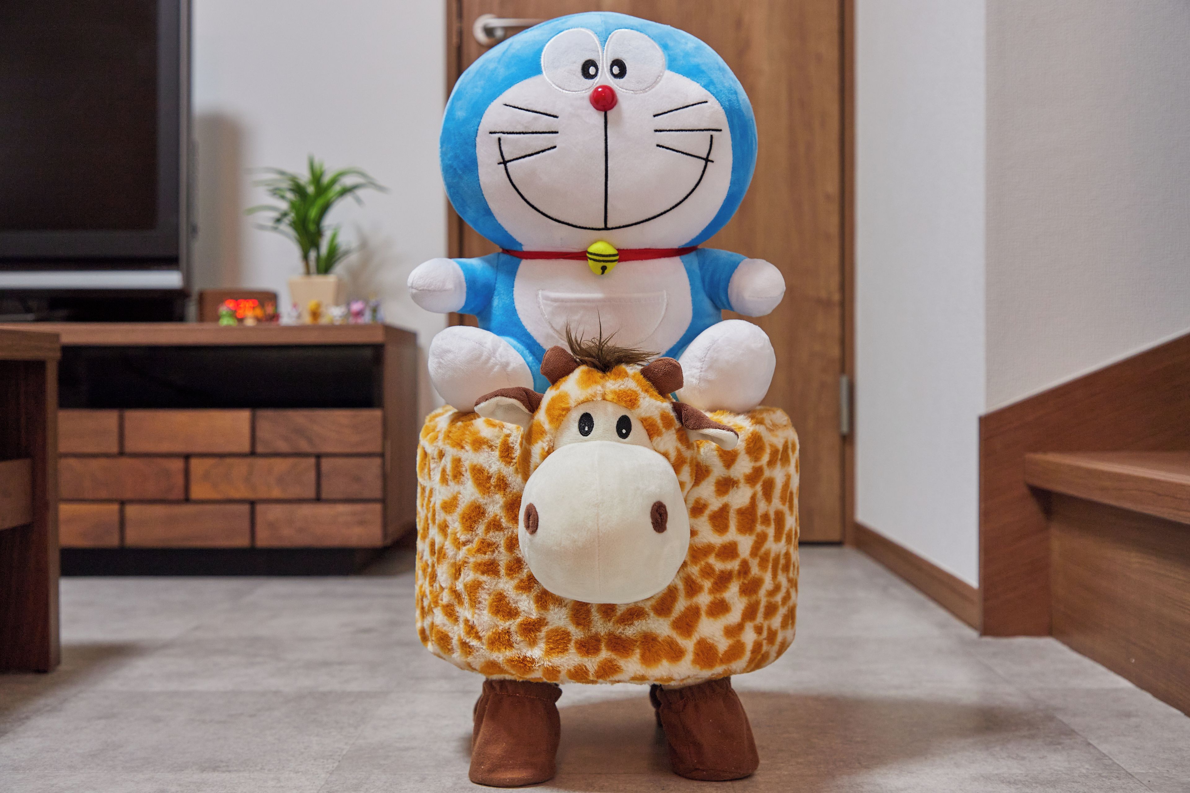 Un peluche de un personaje azul y blanco con disfraz de jirafa, sobre un piso alfombrado.