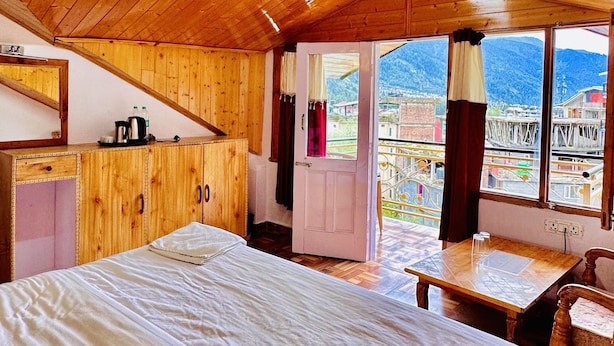A wooden cabin with a large bed, a small table, and a view of a town through a window. - Royal Selection (Manali)