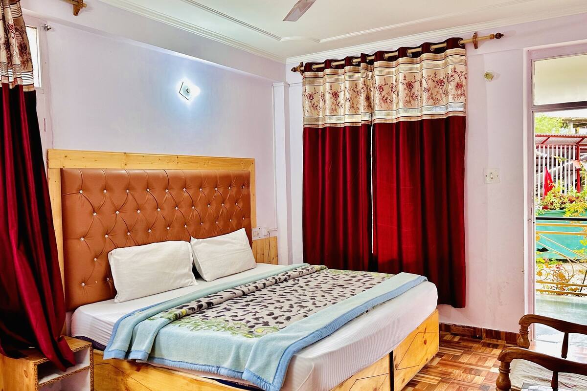 A bedroom with a wooden headboard, a bed with white bedding, red curtains, and a view of a balcony with plants.
