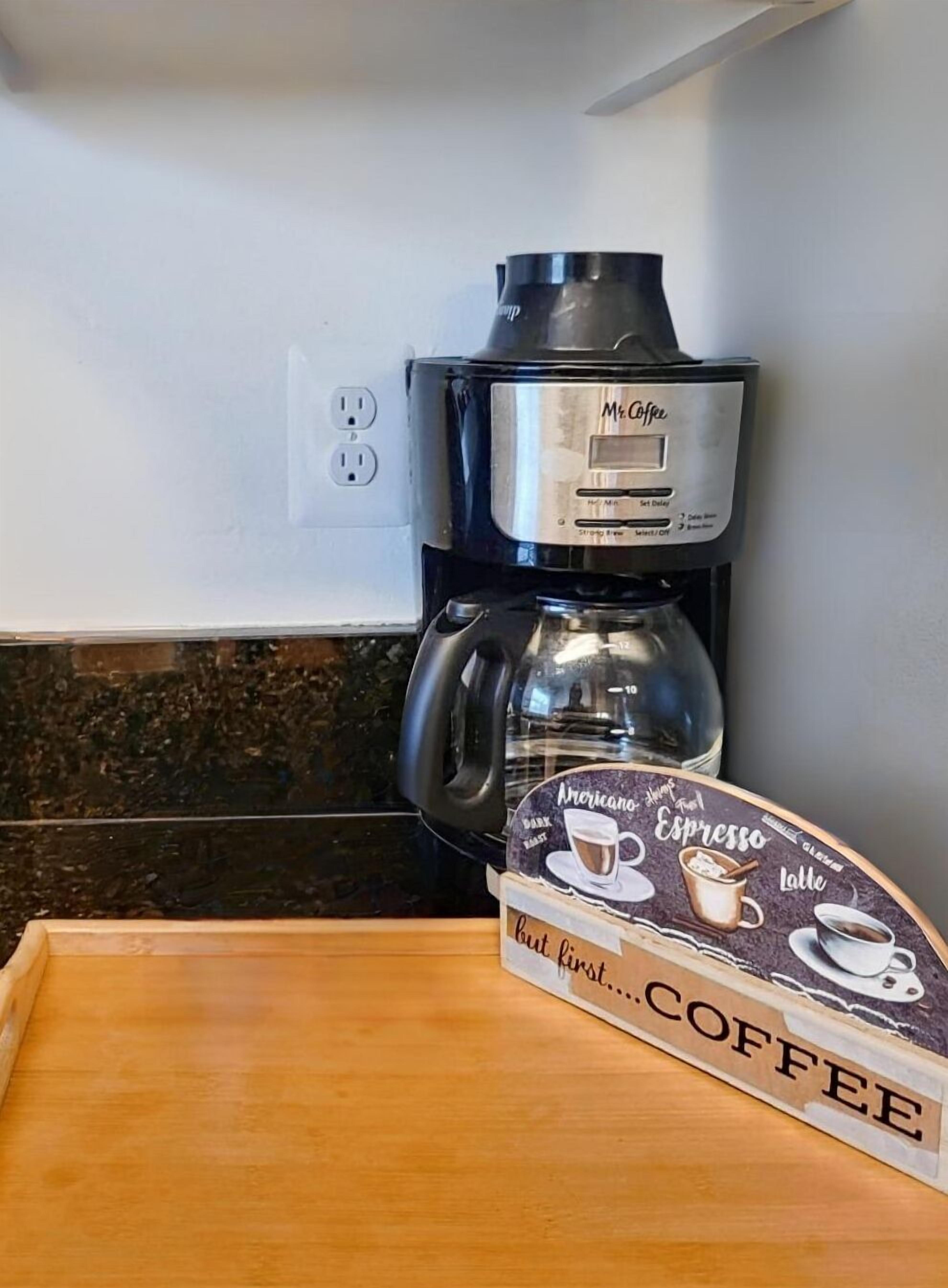 A Mr. Coffee espresso maker on a kitchen counter with a coffee bag labeled "Americano, Espresso, Latte" and the text "but first... COFFEE".