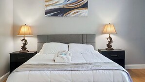 En pænt redt seng med gråt polstret sengegavl og sengefod, med to sengeborde og lamper på hver side.