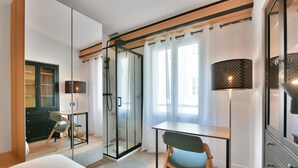 Habitación de hotel moderna con cabina de ducha de vidrio, escritorio con silla, cama y un espejo grande.