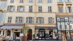 Un edificio con varias ventanas y contraventanas, una pizzería llamada "Le Dauphin" con mesas al aire libre, y un cartel que dice "ZOA".