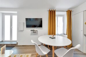 A modern room with a white round table, white chairs, a flat-screen TV mounted on the wall, and a window with yellow curtains.