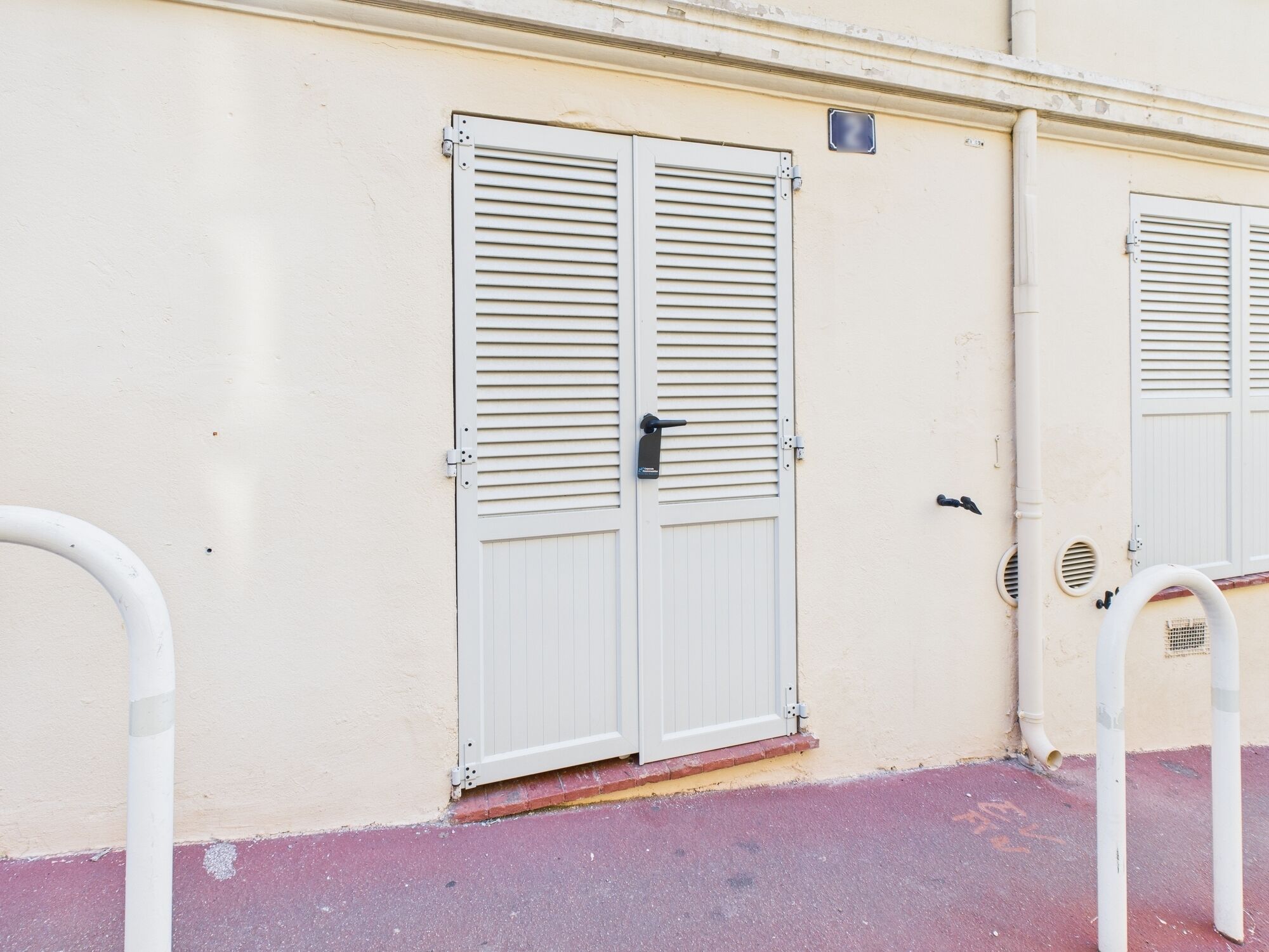 Un edificio con dos puertas blancas y contraventanas horizontales, una pequeña señal sobre la puerta izquierda y una tubería metálica en el suelo.