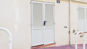 A building with two white doors with horizontal louvered shutters, a small sign above the left door, and a metal pipe on the ground. - Le Palme D'or près de la Croisette et Palais (Cannes)