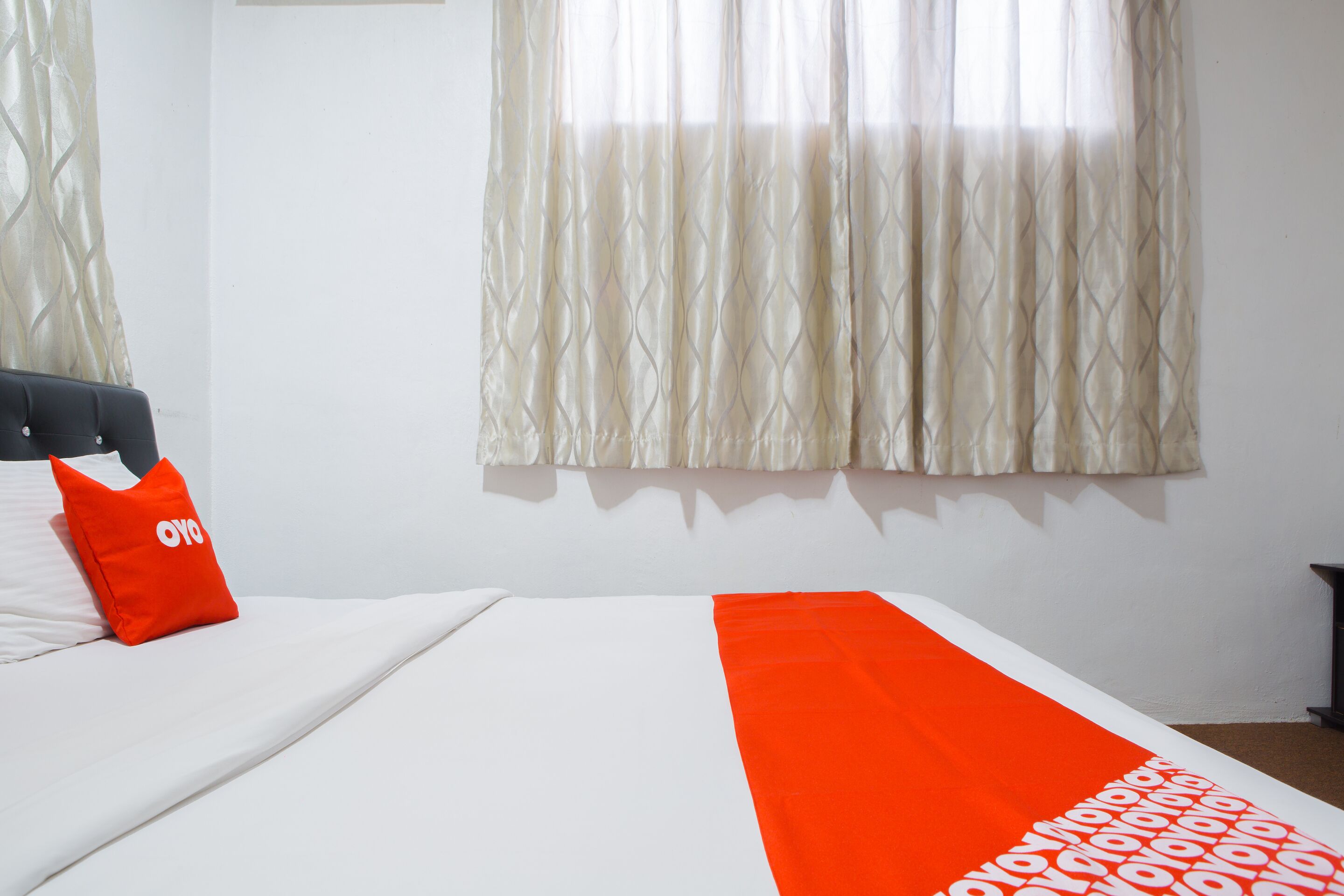 Un letto con copriletto bianco e arancione, un cuscino rosso con un logo e una finestra con tende a motivi.