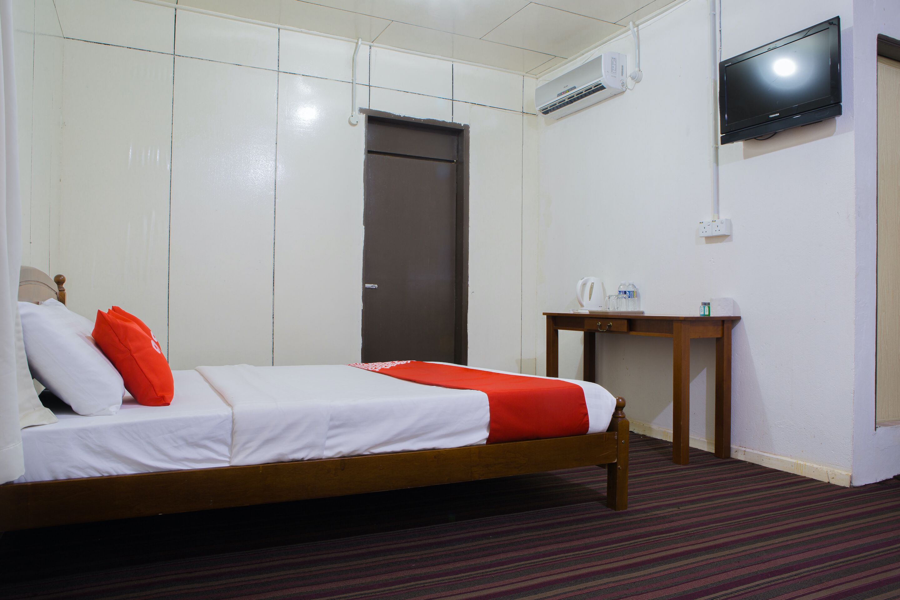 A hotel room with a bed, a wooden nightstand, a television mounted on the wall, and a door.