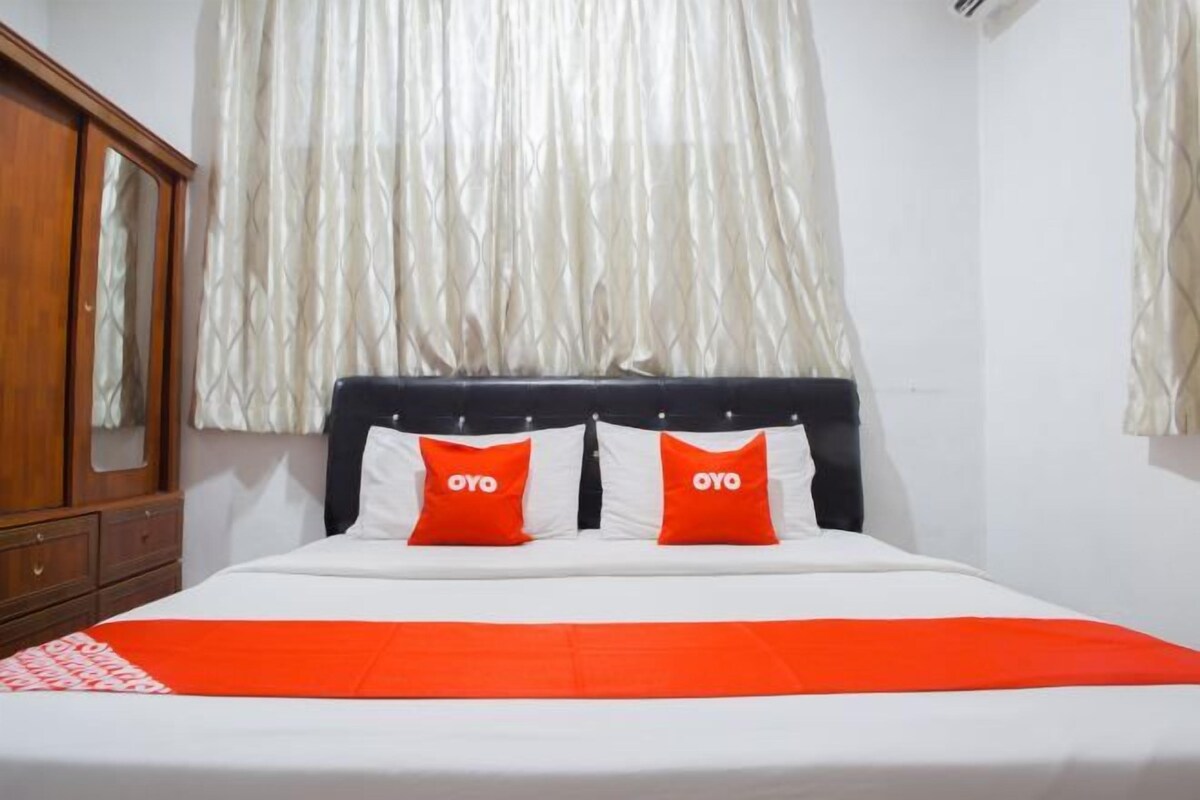 Una cama con ropa blanca y detalles naranja, un armario de madera y una ventana con cortinas traslúcidas.