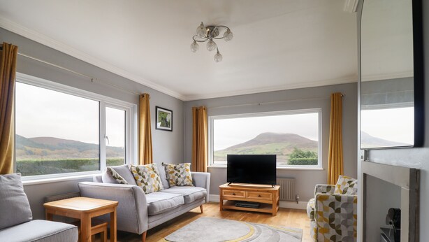 A modern living room with a sofa, a coffee table, and a TV. There are large windows with curtains, a fireplace, and a view of hills. - Craig Yr Ronwy (Bala)