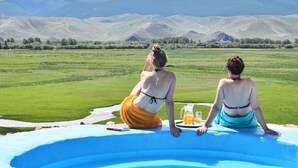 Deux personnes sont assises au bord d’une piscine, avec vue sur des champs verdoyants et des montagnes.
