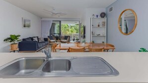 A modern kitchen with a sink, dining area with wooden furniture, a sofa, and a view of greenery outside. - 3 Bedroom Poolside Drift North Apartment (Tweed)