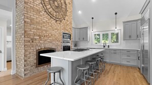 A modern kitchen with a brick fireplace, stainless steel appliances, and a large island with bar stools. - Waterfront, Pool & Dock | Shoreway Cove by AvantStay (Huntersville)