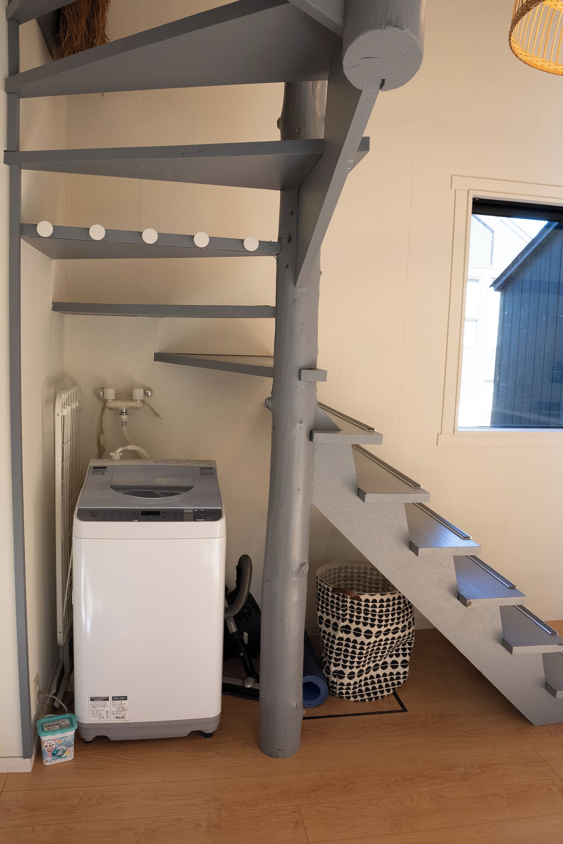 A compact room with a grey spiral staircase, a white washing machine, and a window with a view of the outside.