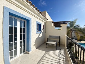 Een balkon met een betegelde vloer, een bankje, een zwembad en een gebouw met blauwe accenten en een witte muur.