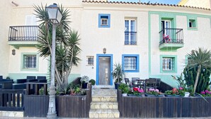 Um edifício com porta azul, várias janelas e varandas. Possui áreas de estar ao ar livre e vasos com plantas.