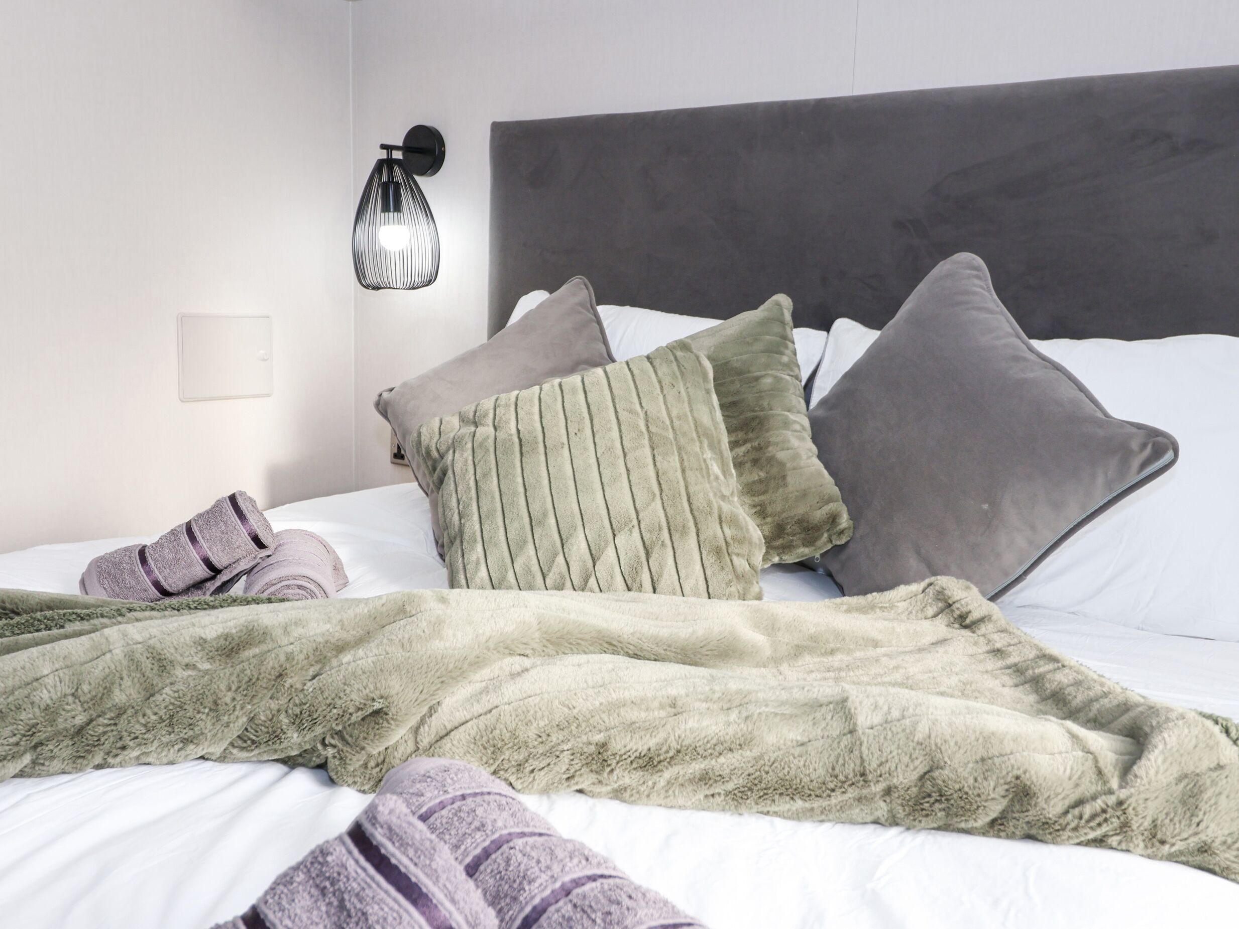 Un lit bien fait, avec une tête de lit grise, une couverture verte et moelleuse, et plusieurs oreillers dans différentes nuances de gris et de vert.
