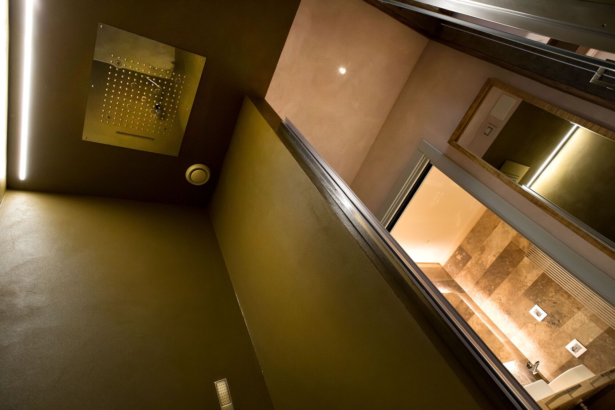 Un intérieur moderne avec un panneau métallique, un luminaire encastré au plafond et une vue sur une salle de bain équipée d’un lavabo et d’un miroir.