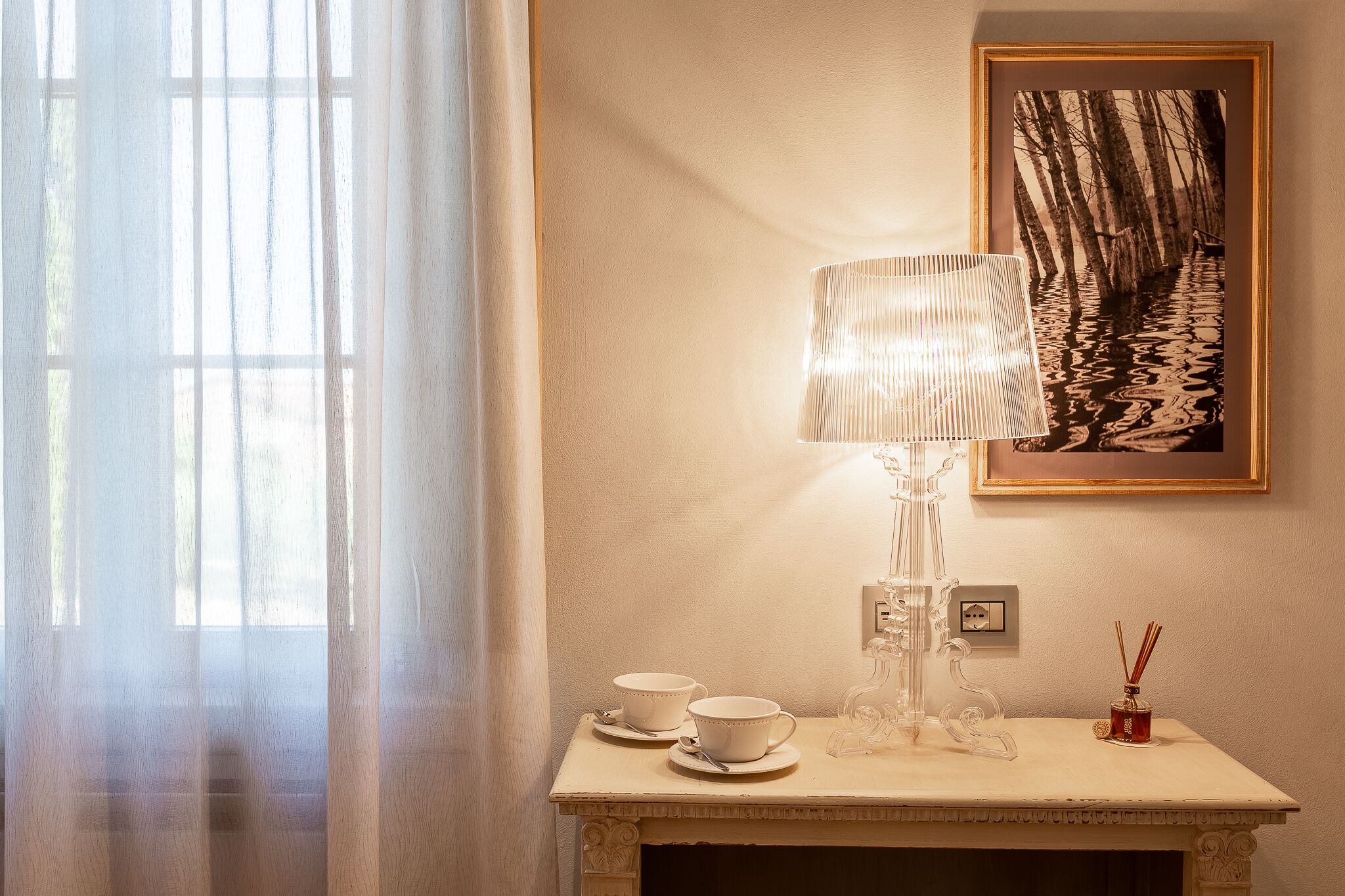 Una stanza con un tavolo, due tazze, una lampada, un quadro incorniciato e una finestra con tende leggere.