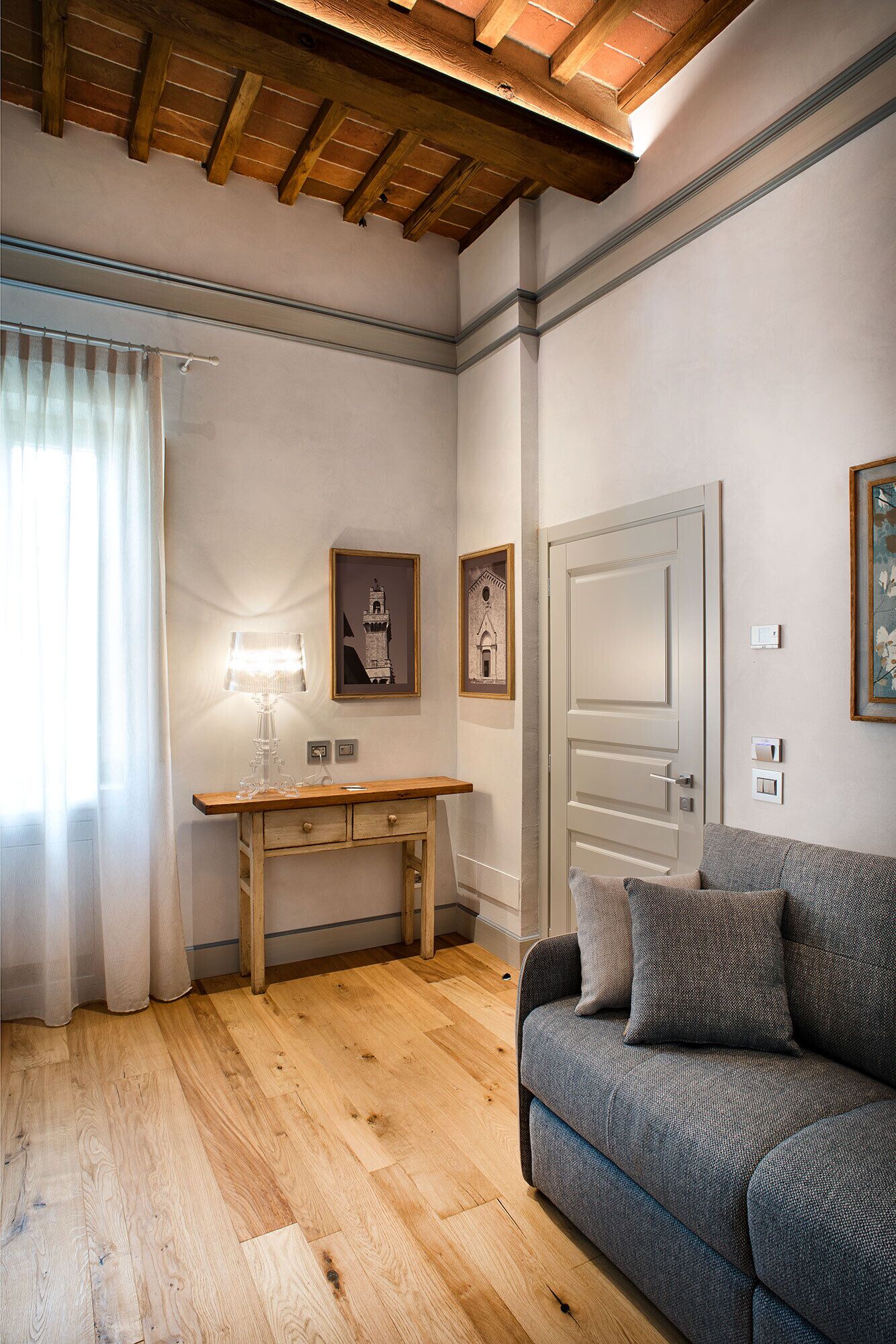 Une pièce avec un bureau en bois, une lampe, un canapé gris et des tableaux encadrés accrochés au mur.