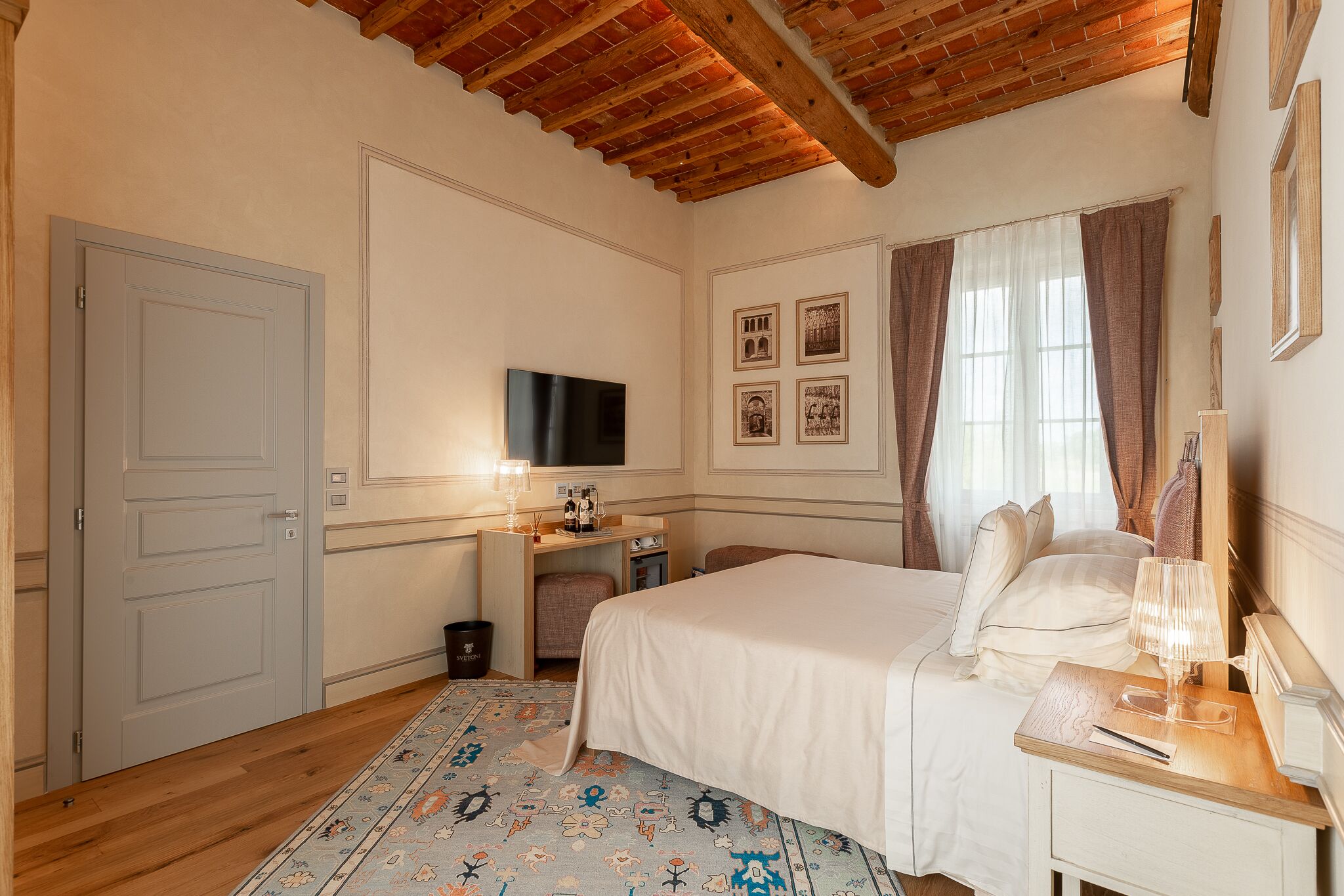 Habitación de hotel con techo de madera, cama con ropa blanca, escritorio con lámpara, televisor y cuadros en la pared.