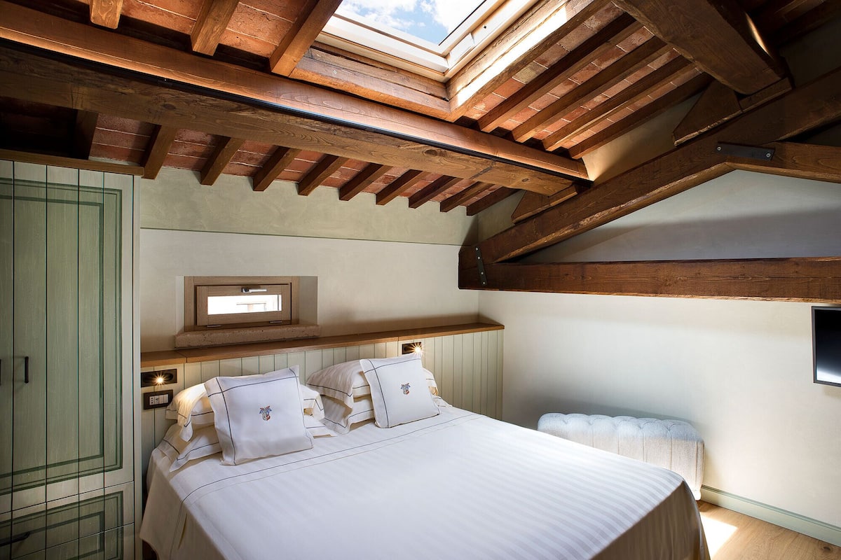 A bedroom with a skylight, wooden beams, a bed with white linens, a bedside table, and a small window.