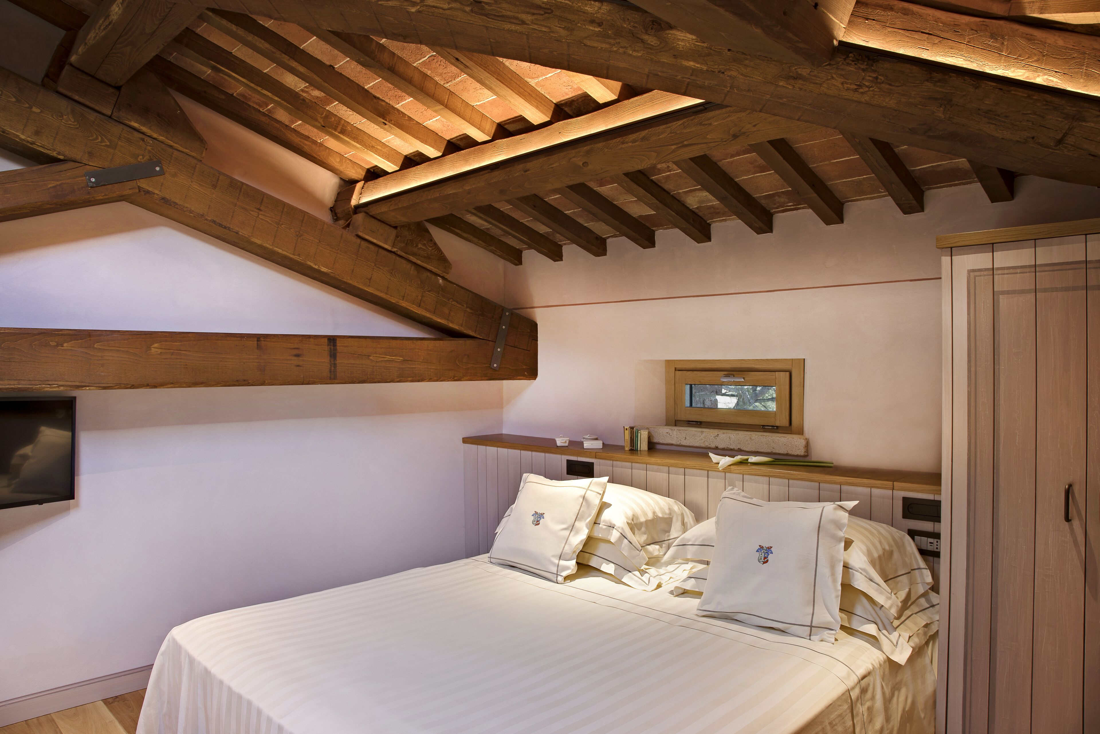 Un dormitorio con techo de madera, una cama con ropa blanca, una mesita de noche, un televisor montado en la pared y un armario.