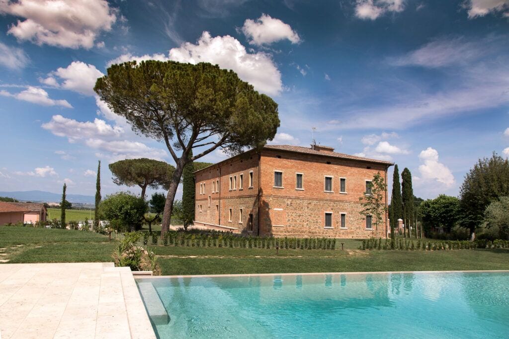 A brick building with a swimming pool in front, surrounded by greenery and trees.