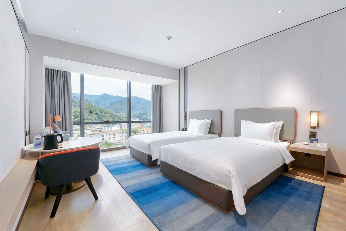 Quarto de hotel com duas camas, uma escrivaninha, uma cadeira e uma janela grande com vista para as montanhas.